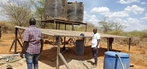 Two men stand near a makeshift shade structure with large water tanks elevated on a wooden platform in the dry, arid environment. The sky is clear and blue. One man faces the camera while the other has his back turned. Various pipes and containers, part of a recent water donation effort, are nearby.