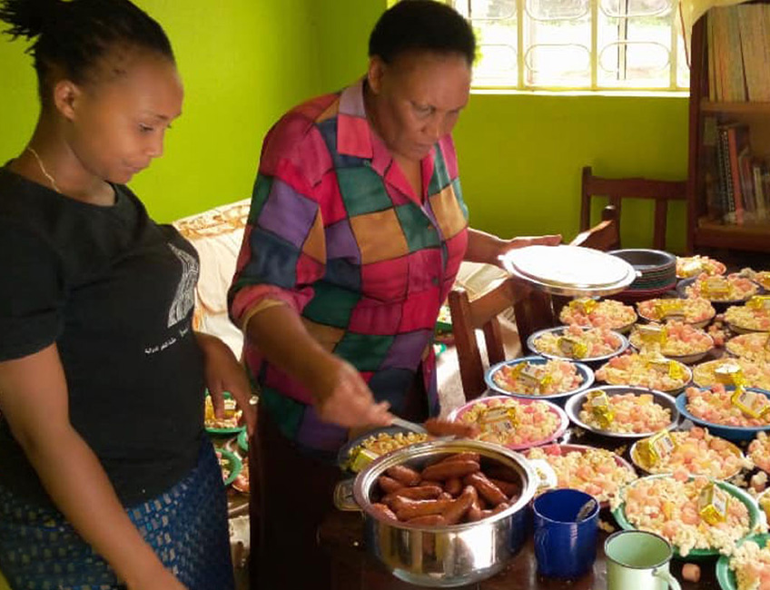 Two women are preparing a large number of plates with food in a brightly lit room. One woman, wearing a multi-colored top, and the other in a black shirt, are setting up for a charity event. The table is covered with plates filled with diverse food items, and a pot of sausages is central.