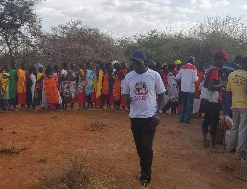 A group of people, many wearing brightly colored traditional clothing, are gathered outside in a rural area with sparse vegetation. A man in a white T-shirt and blue cap walks in the foreground, likely discussing a recent water charity donation, while others stand and converse in the background.