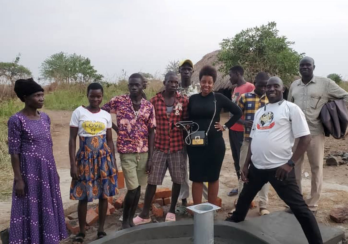 A group of nine people stand around a newly constructed well in an outdoor rural setting, their smiles radiating the joy of providing water through charity. The group includes men and women in casual clothing, and they are beaming at the camera. Trees and a rocky hill can be seen in the background.